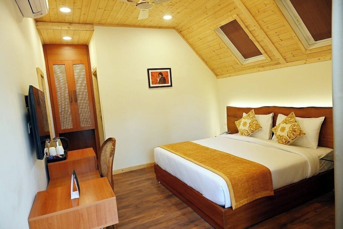 Een hotelkamer met een houten plafond, een bed met wit en goud linnengoed, een houten bureau met een stoel, en een ingelijst schilderij aan de muur.