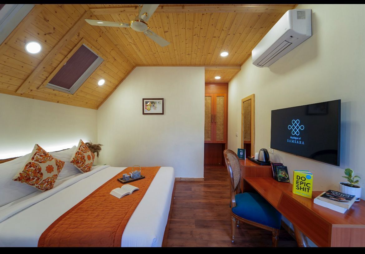 A hotel room with a wooden ceiling, a bed with a white and orange bedspread, a desk with a chair, a flat-screen TV, and a small potted plant.