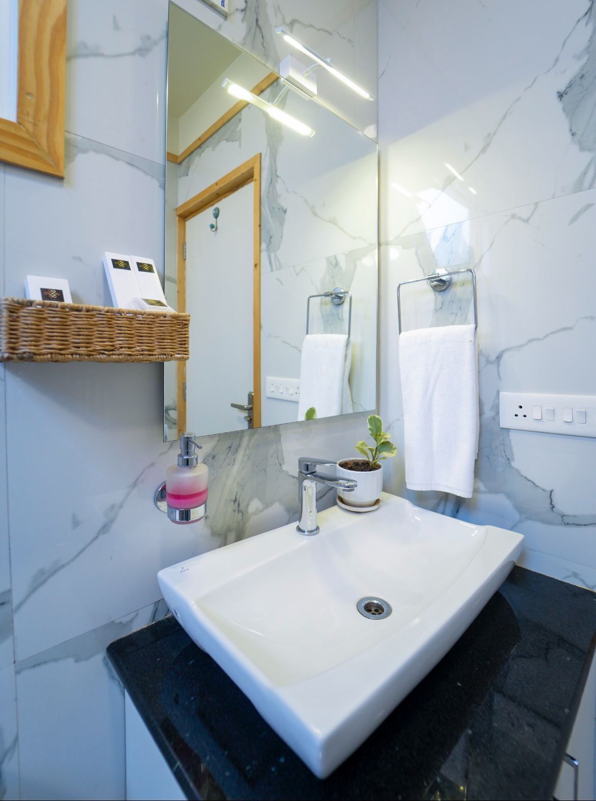 A modern bathroom with a white marble sink, a mirror, a small potted plant, and a wicker basket with toiletries.