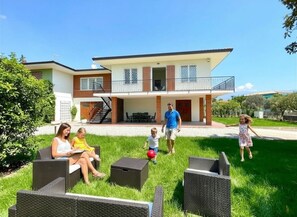 Eine Familie mit Kindern spielt auf einer Rasenfläche vor einem zweistöckigen Haus mit Balkon.