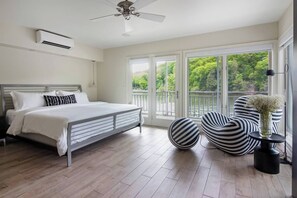 A bedroom with a bed, a ceiling fan, a large window with a view of greenery, and striped chairs.