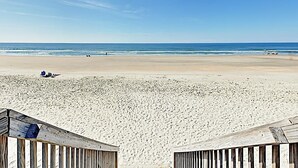Une plage avec une promenade en bois menant à l’eau, quelques personnes visibles au loin et un ciel dégagé.