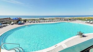 Une grande piscine à eau claire, dotée de chaises longues et de parasols, et entourée d’une terrasse offrant une vue sur la plage.