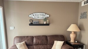 Un sofá de cuero marrón con dos cojines, un letrero en la pared que dice "WELCOME TO THE LAKE", y una mesita con una lámpara y objetos decorativos.