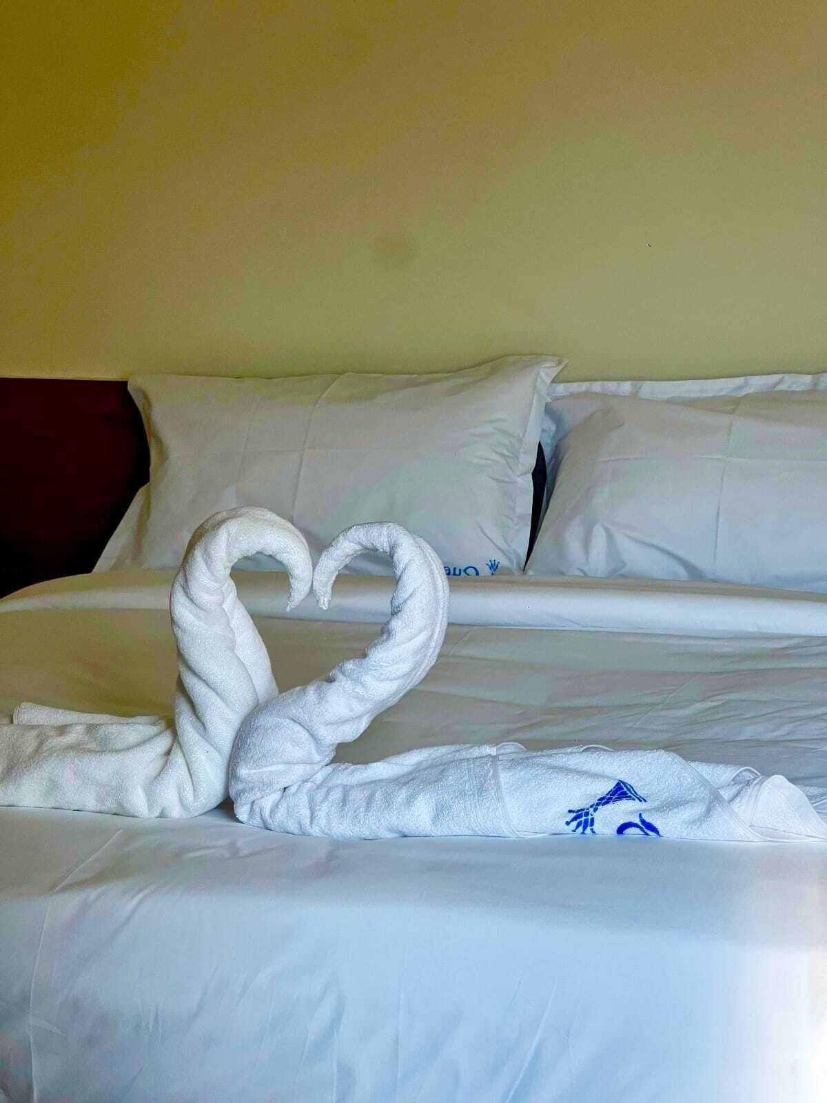 A hotel room with a neatly made bed, white linens, and a heart-shaped towel decoration on the bed.
