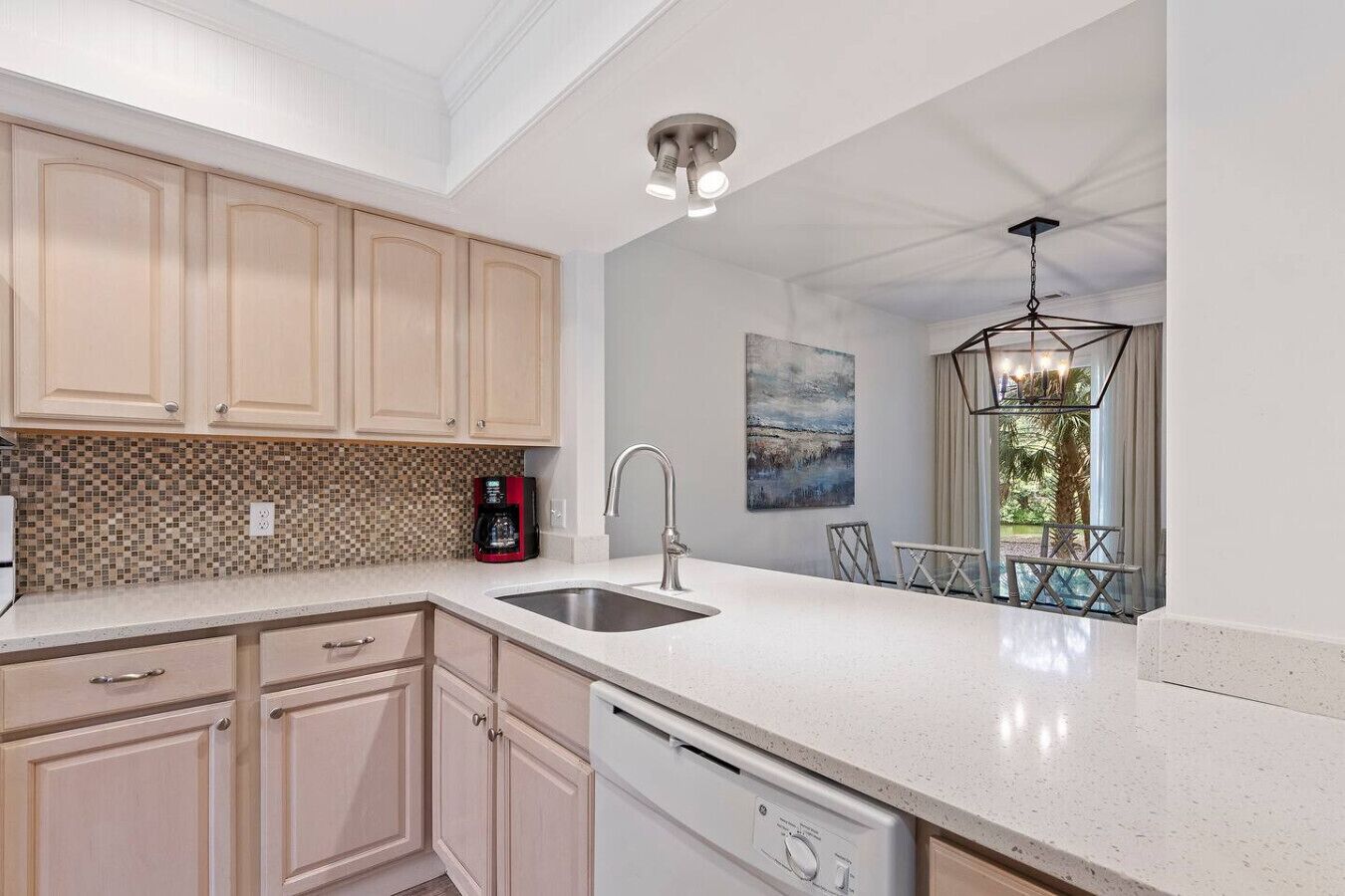 A modern kitchen with light wood cabinets, a white countertop, a built-in dishwasher, and a coffee maker.