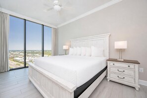 A bedroom with a large bed, a nightstand, a lamp, and a view of a cityscape through large windows. - New Luxury Gulf Front Phoenix Gulf Shores II 1501 (Gulf Shores)
