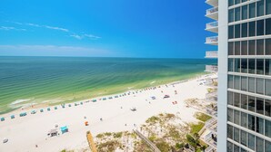 A beach with a clear view of the ocean, a white building with balconies, and a sandy beach with umbrellas.