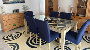 Un comedor con mesa de vidrio, sillas tapizadas en azul, un mueble de madera con puertas de cristal y un cuadro de veleros en la pared.