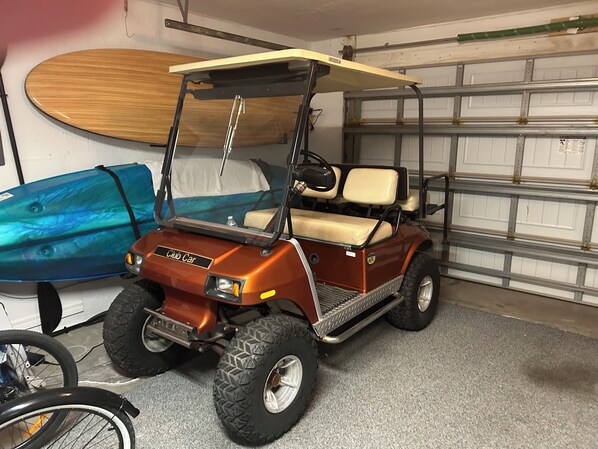 En golfbil parkeret i et garage med et surfbræt og en cykel i nærheden.