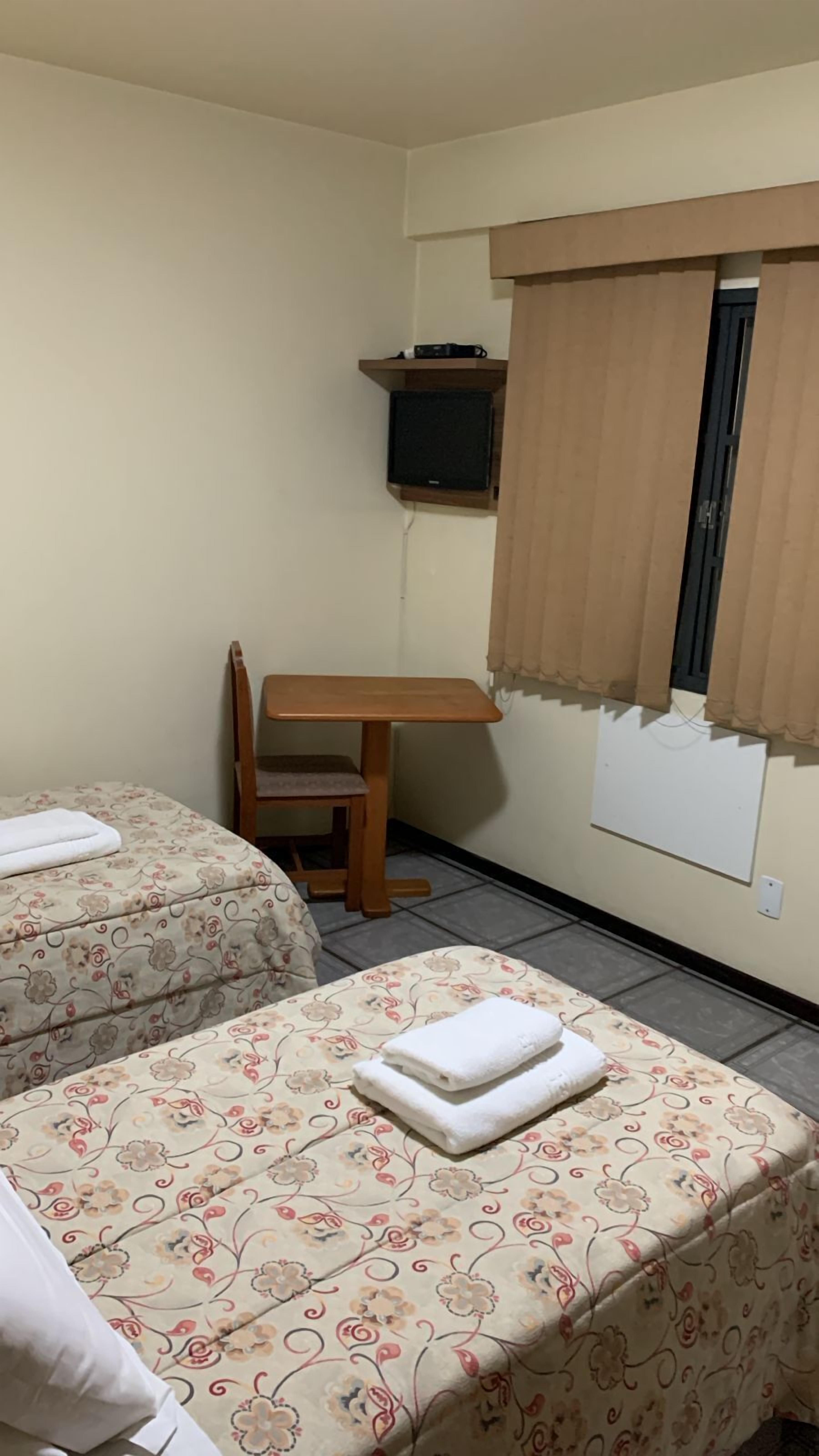 Una habitación pequeña y sencilla con dos camas, un escritorio de madera, una silla y un televisor sobre una estantería.