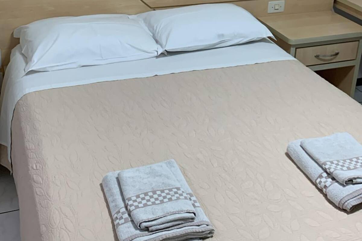 Una cama bien hecha con una colcha color beige y dos toallas dobladas, una mesita de noche de madera con una lámpara, y dos cuadros en la pared.