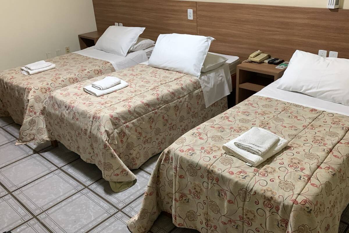Habitación de hotel con dos camas individuales, cada una con cubrecama floral y una toalla doblada. Se observa un cabecero de madera, una mesita de noche con teléfono y una lámpara fijada a la pared.