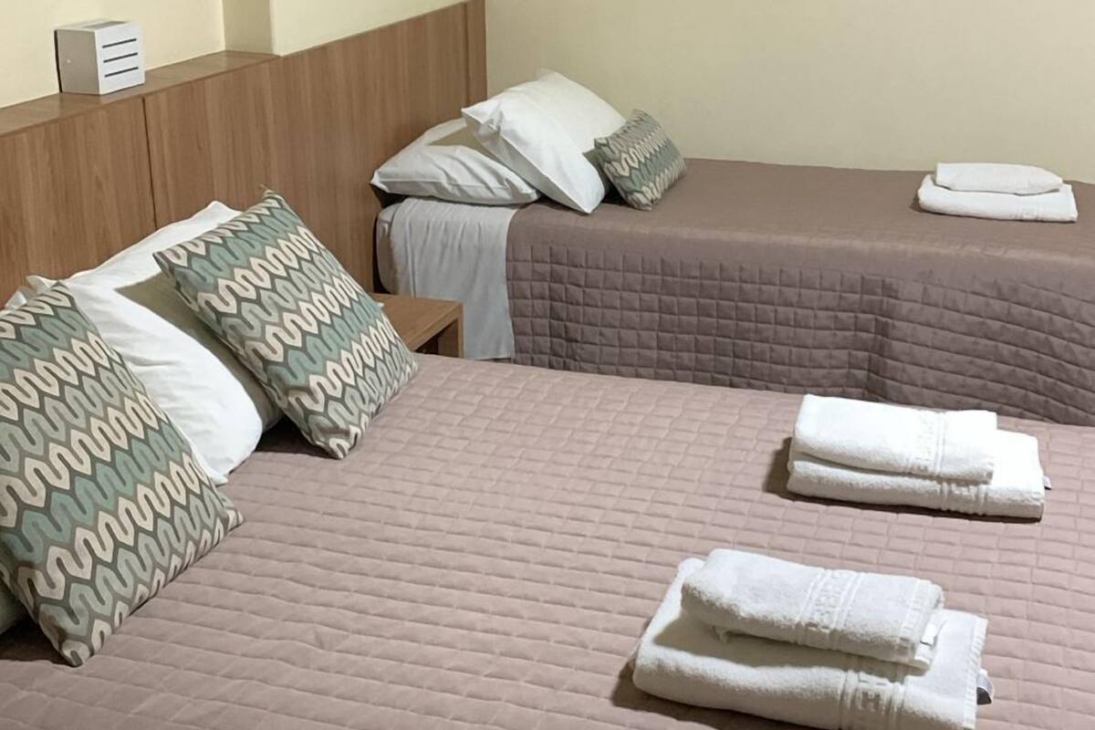 Habitación de hotel con dos camas, cada una con una colcha acolchada y almohadas. Hay toallas sobre las camas y una mesita con una caja encima.