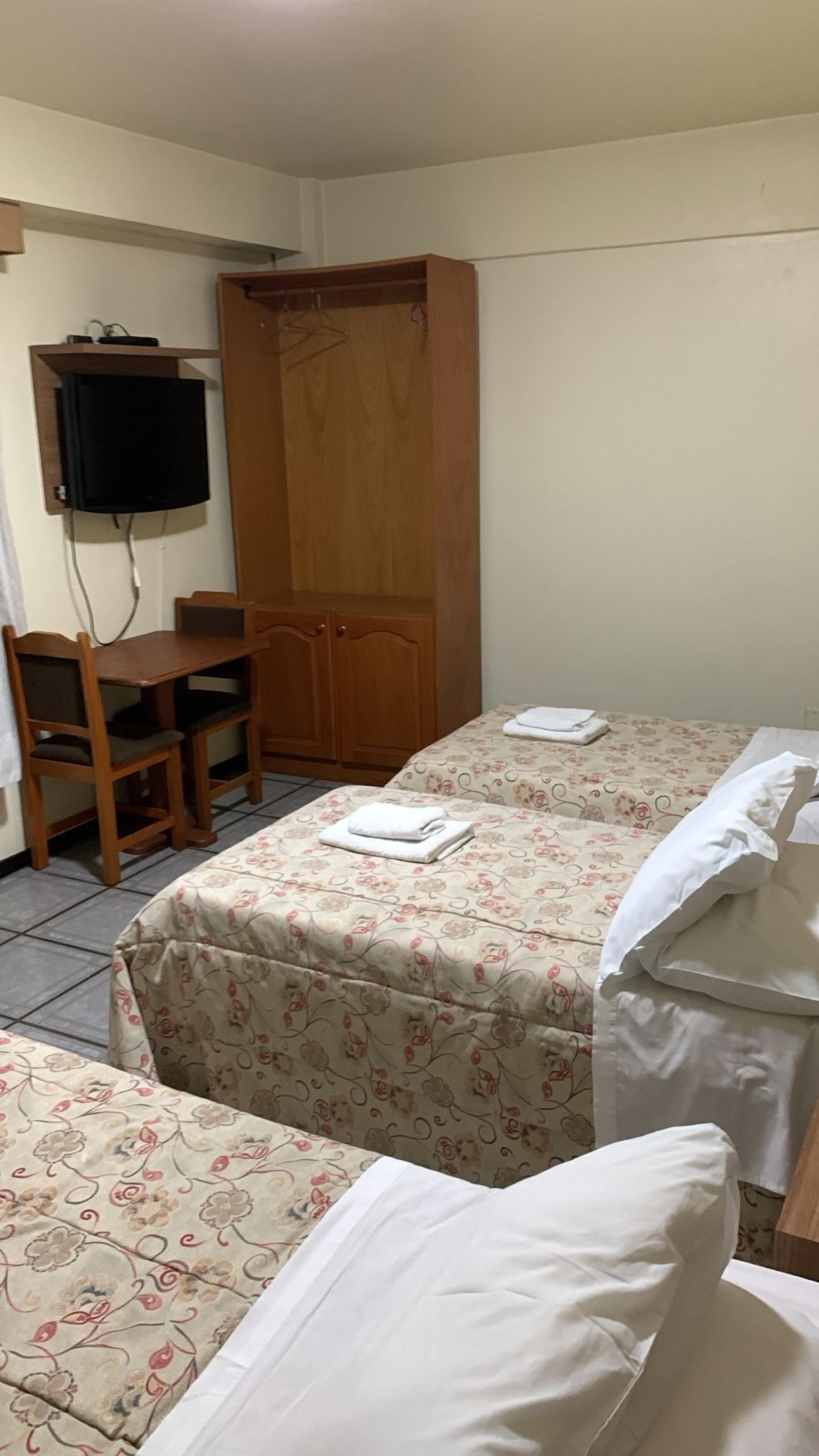 A small, tidy room with two single beds, a wooden desk with a chair, a TV mounted on the wall, and a wardrobe.
