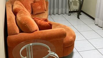 Una sala de estar con un sofá naranja grande, una mesa de centro de vidrio, una lámpara y una cortina.