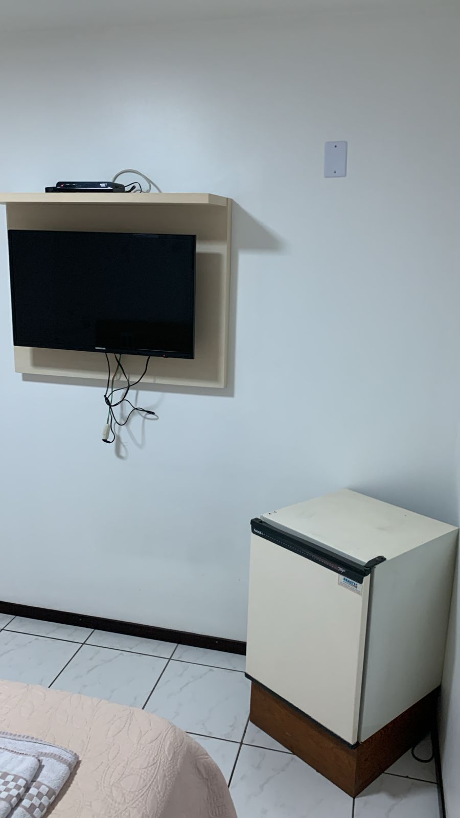 Una habitación con un televisor montado en la pared, un refrigerador pequeño y una cama con colcha estampada.