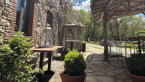 Un patio exterior con suelo de piedra, una mesa de madera, plantas en macetas, un farol y una estructura tipo pérgola.