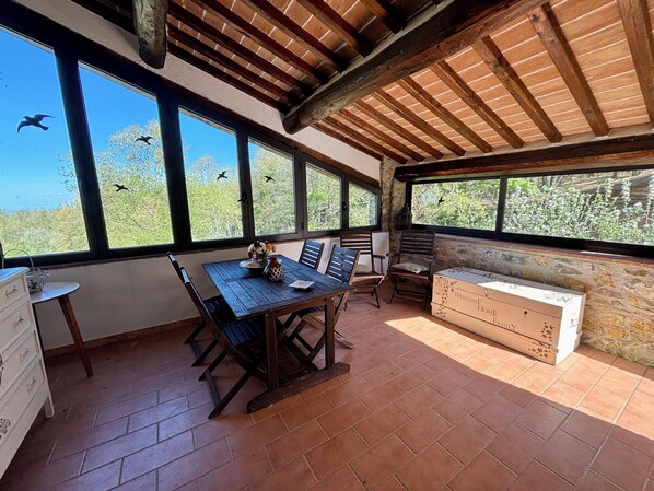 Un comedor rústico con una mesa y sillas de madera, una pared de piedra y ventanas amplias con vistas a árboles y pájaros.