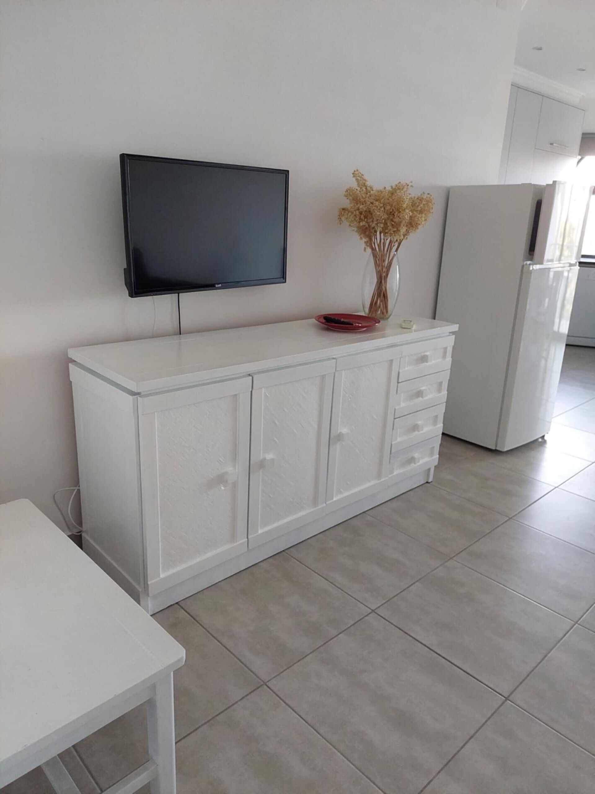 A modern kitchen with a white cabinet, a flat-screen TV mounted above it, and a refrigerator to the right.