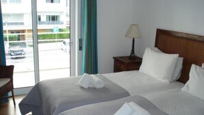 Habitación de hotel con dos camas, una mesita de noche con lámpara, una silla y vistas al exterior a través de un ventanal.