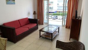 Una sala de estar con un sofá rojo, una mesa de centro de vidrio y una silla de mimbre. Hay una lámpara y un cuadro en la pared.