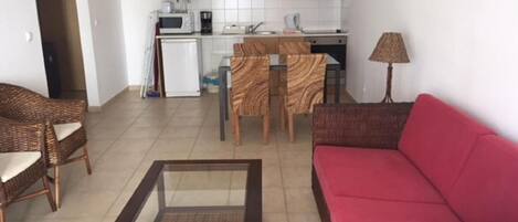 A compact living space with a kitchenette, a red sofa, wicker furniture, and a glass-top coffee table.