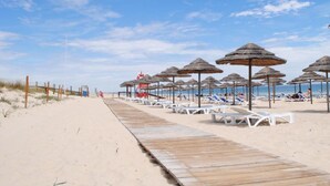 Ein hölzerner Steg führt zu Strandliegen und Sonnenschirmen am Sandstrand.
