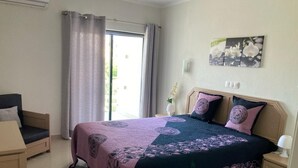 Ein Schlafzimmer mit Bett, Stuhl, Nachttisch, einem Fenster mit Vorhängen und einem Wandbild.