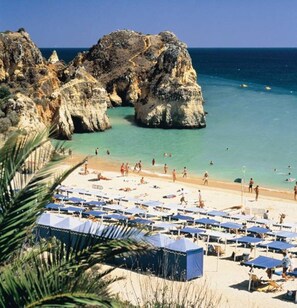 Ein Strand mit felsigen Klippen, klarem türkisblauem Wasser und vielen Strandsonnenschirmen.