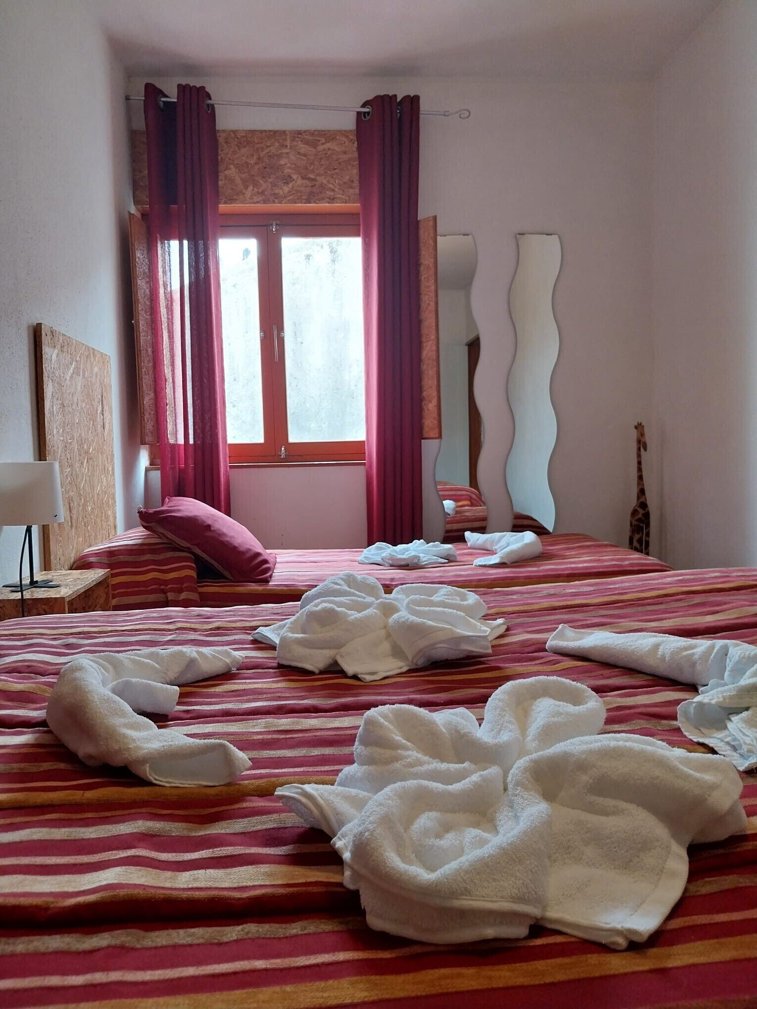 Une chambre d’hôtel avec un lit, des oreillers, une fenêtre avec des rideaux, un miroir et un objet de décoration représentant une girafe.
