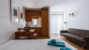 A hotel room with a bed, a sofa, a dining table with chairs, a kitchen area, and a window with curtains.