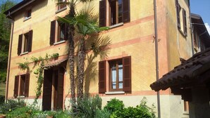 A two-story building with a yellow facade and brown shutters, surrounded by a well-maintained garden with various plants and a palm tree.