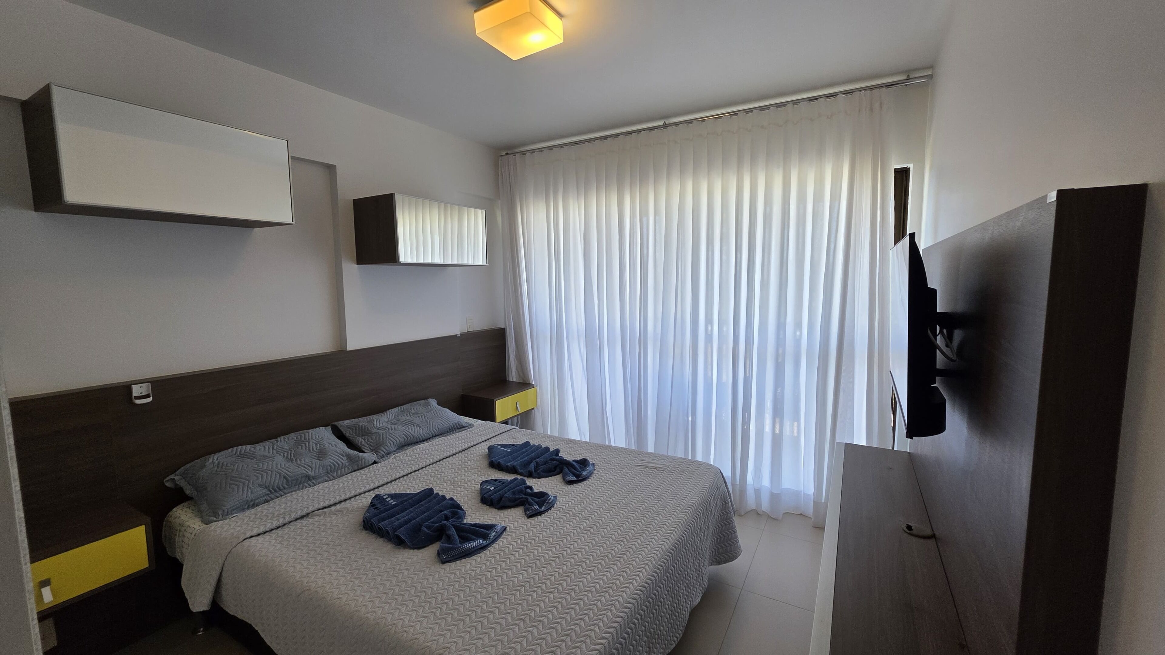 A hotel room with a bed, a bedside table, a TV mounted on the wall, and a window with curtains.