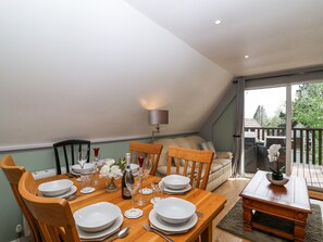 Un coin repas avec une table en bois dressée pour quatre personnes, entourée de chaises en bois. On aperçoit un canapé et une petite table en bois avec une plante au fond.
