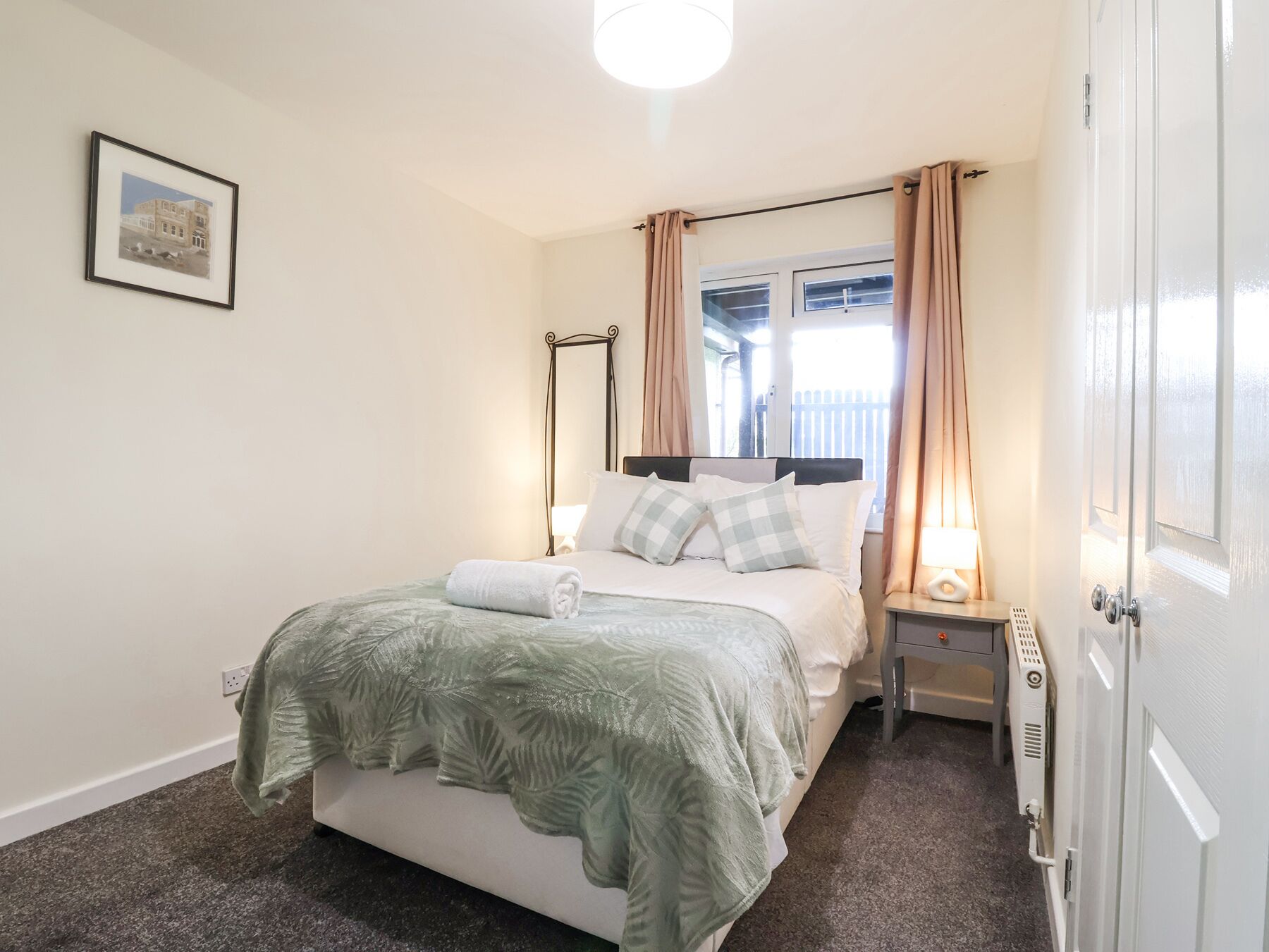 A neatly arranged bedroom with a bed, bedside tables, a window with curtains, a radiator, and a wall-mounted picture.