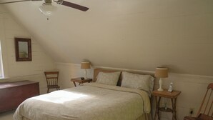 Une chambre à coucher avec un grand lit, deux tables de chevet avec des lampes, un fauteuil à bascule en bois, un bureau en bois et un ventilateur de plafond.