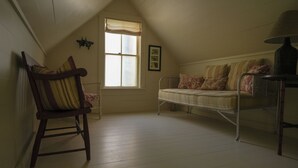 Une chambre mansardée chaleureuse avec un plafond en pente, une fenêtre agrémentée d’une lambrequin, un fauteuil à bascule en bois, un banc rembourré et une lampe.