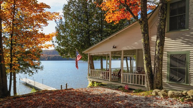 Un chalet au bord d’un lac, doté d’une véranda donnant sur l’étendue d’eau, avec un drapeau américain et un feuillage d’automne.