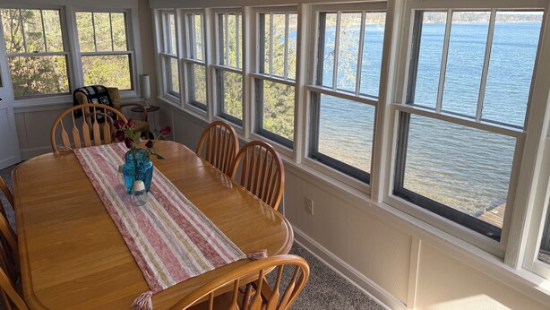 A dining area with a wooden table, chairs, and a view of the water through large windows. - Retro Lakeside Charmer in Wonderful Wautoma with WiFi, AC and lake frontage (Wautoma)
