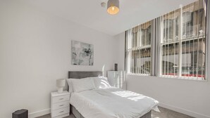 A bedroom with a bed, a bedside table, a lamp, a small cabinet, and a window with blinds. - GuestReady - Urban getaway in Liverpool (Liverpool)