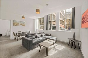 A modern living room with a grey sectional sofa, a small coffee table, and a dining area with a white table and chairs. Large windows offer views of the city. - GuestReady - Urban getaway in Liverpool (Liverpool)