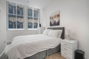 A bedroom with a bed, a nightstand, a lamp, a picture on the wall, and a window with blinds. - GuestReady – Modern comfort in Liverpool (Liverpool)