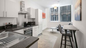 A modern kitchen with white cabinets, a built-in oven, and a microwave. There is a dining area with a table and chairs, and a window with a view of the city. - GuestReady – Modern comfort in Liverpool (Liverpool)