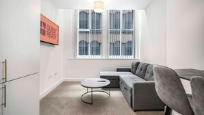 A modern, compact living space with a grey sofa, a small round table, and a window with blinds. - GuestReady – Modern comfort in Liverpool (Liverpool)