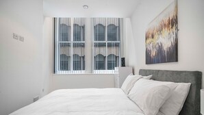 A bedroom with a large bed, a window with blinds, a bedside table, and a wall-mounted painting. - GuestReady – Modern comfort in Liverpool (Liverpool)