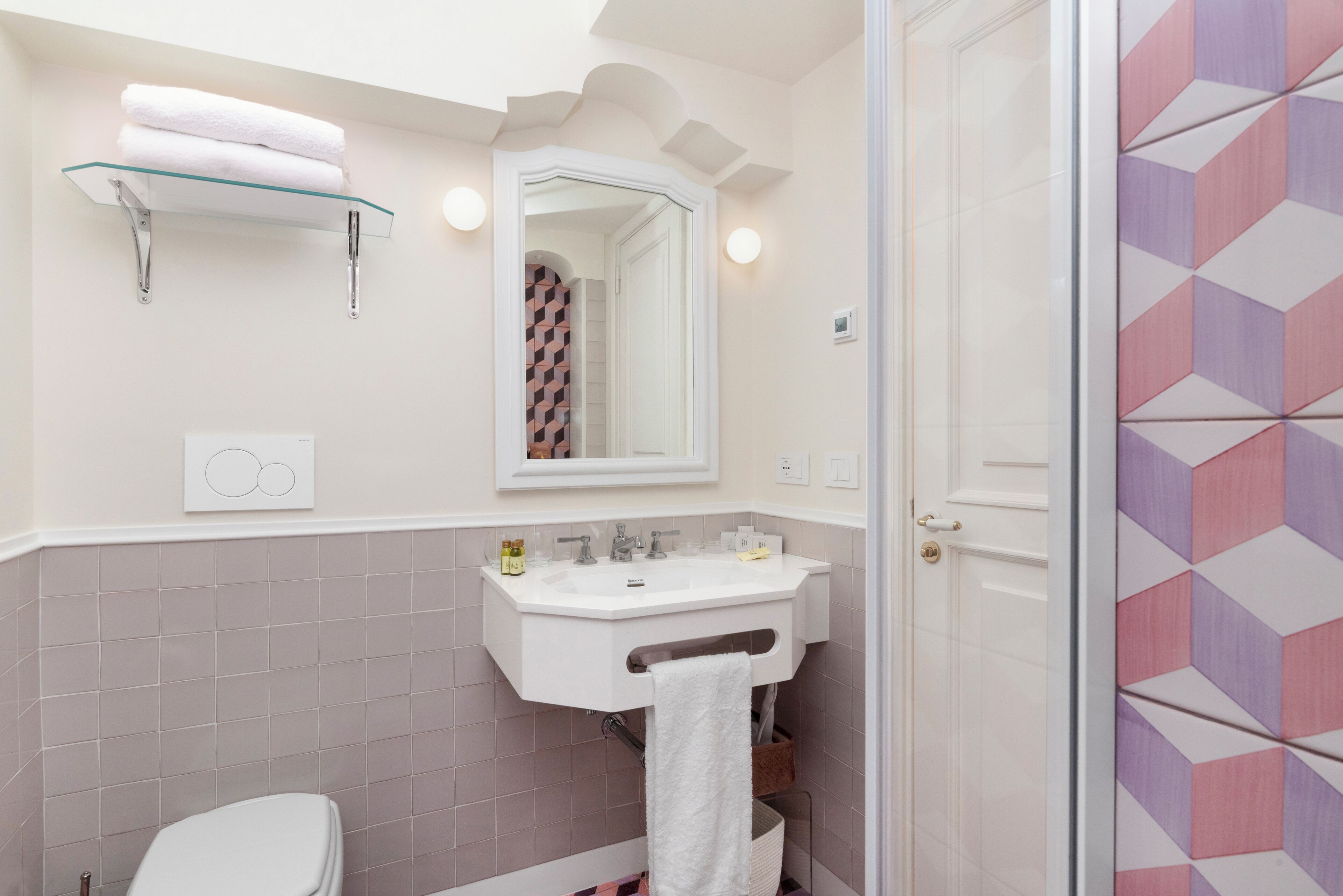 A bathroom with a white sink, a mirror, and a toilet. There is a towel rack with towels and a shower area with a geometric tile pattern.