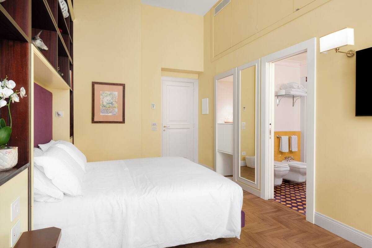A hotel room with a bed, a bedside table, a shelf with decorative items, a framed picture on the wall, and an open door leading to a bathroom.