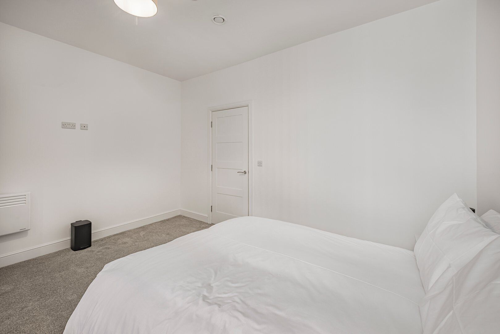 Een witte kamer met een bed, een deur, een verwarming en een prullenbak.