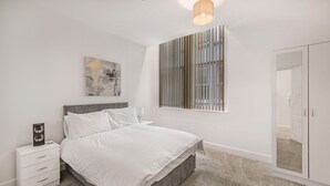 Une chambre à coucher comprenant un lit, des tables de chevet, une commode, une œuvre d’art accrochée au mur, une fenêtre avec des stores et une armoire.